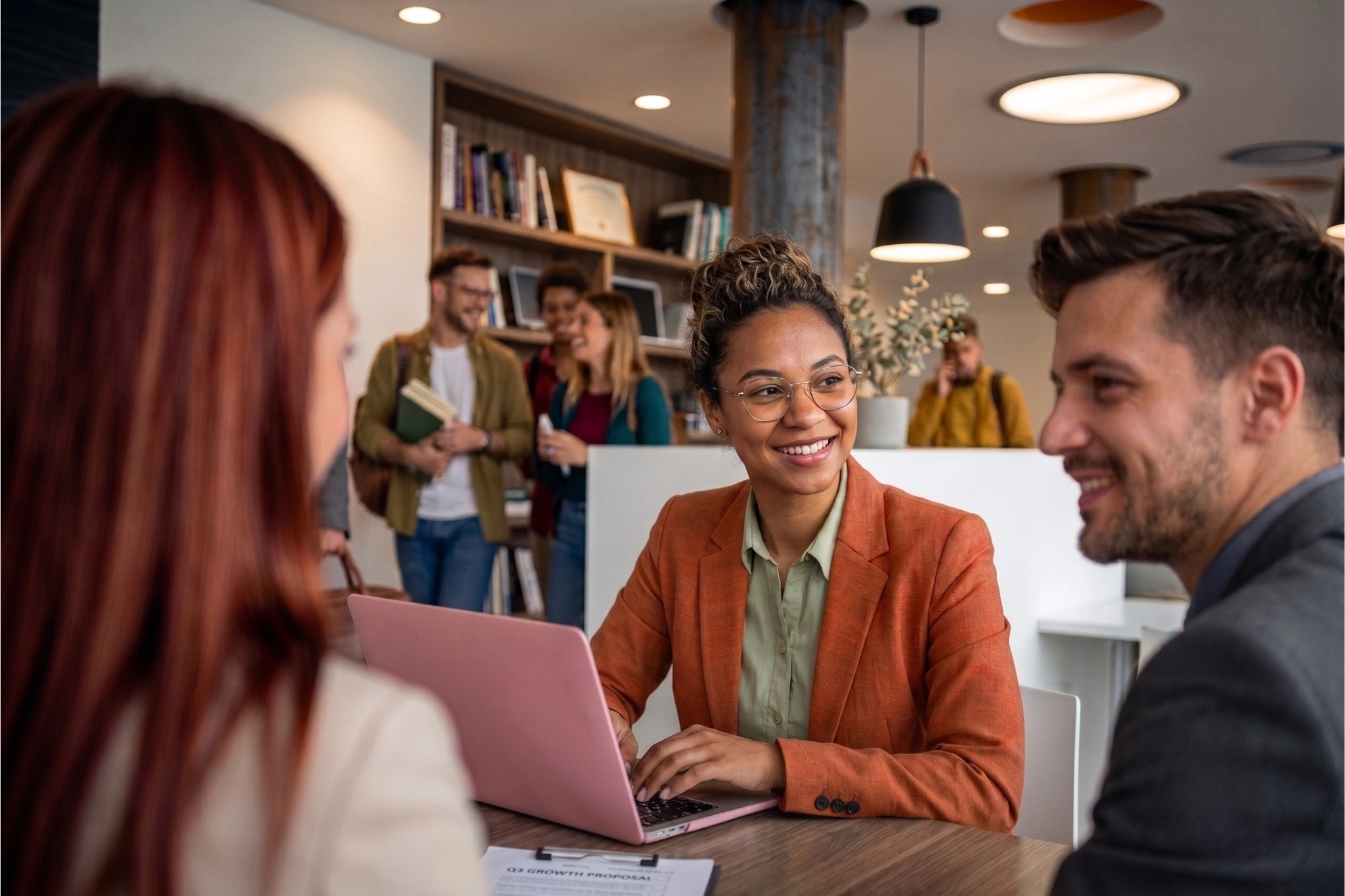 A college admissions counselor smiles while meeting with two prospective students across a table in a modern campus admissions office. A pink laptop and paperwork sit on the table, while other students talk in the background near bookshelves and a reception desk.